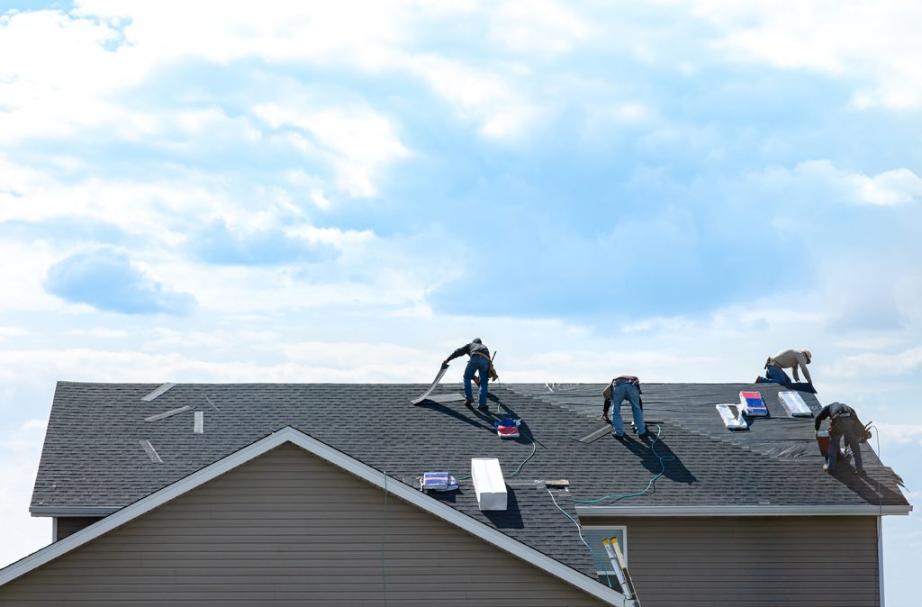 4 construction workers fixing roof against clouds blue sky. Roofer install shingles at the top of the house. Renovate, improve, build home exterior by professional teamwork. Safety, protection concept