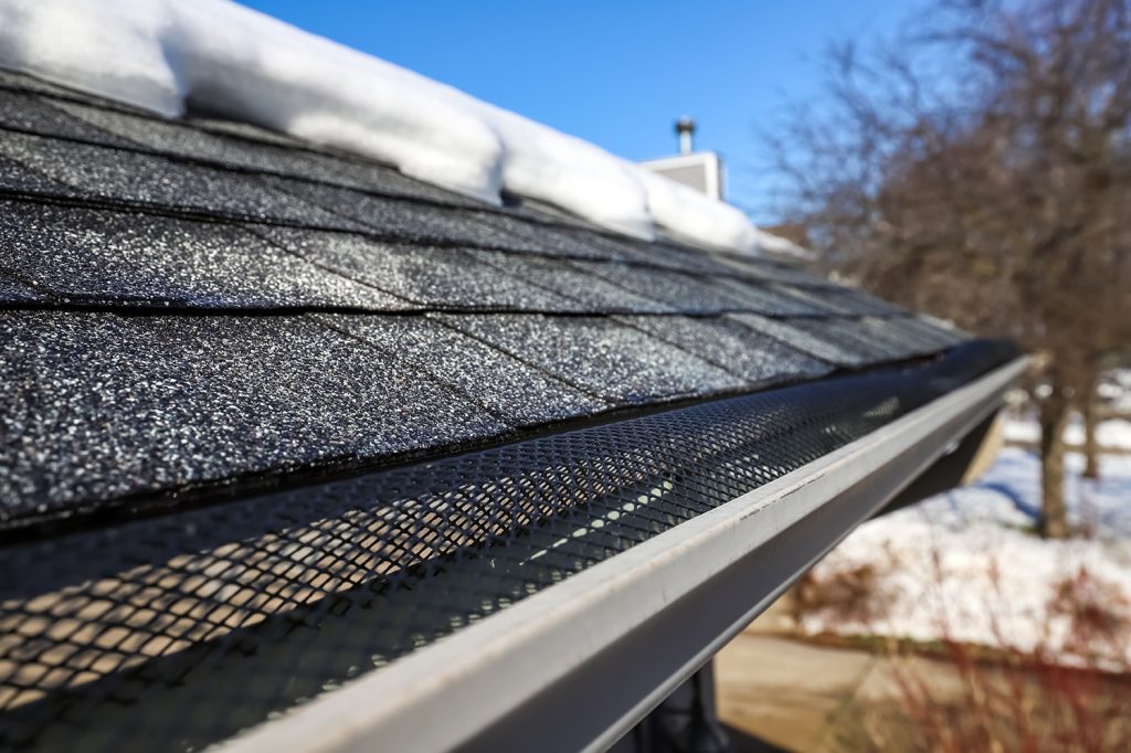 Shallow focus on mesh in gutter and shingles, gutter in winter and snow melted off of roof to prevent roof damage